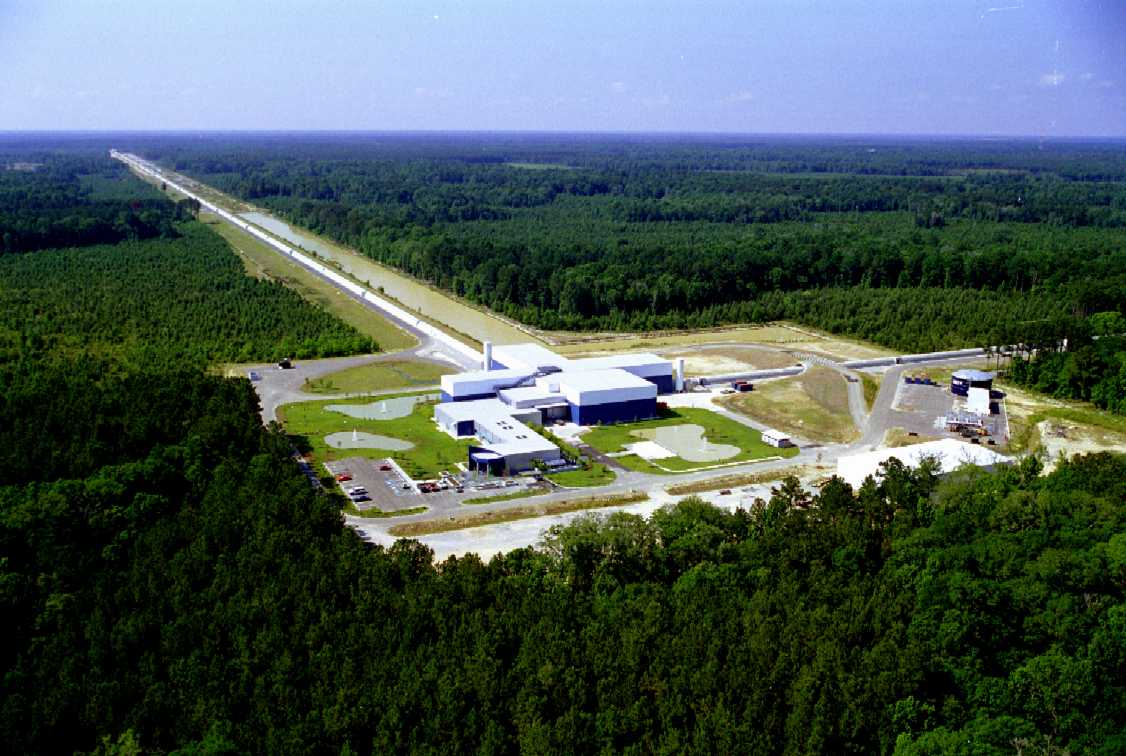 An aerial view of the LIGO instrument in Louisiana. This picture shows the central building, which contains the laser, the semi-silvered mirror, and the detector. We can see the large concrete tubes going out in two directions from the center, which encase the arms securely in large vacuum tubes; one goes off to the right, while the other extends to the top left of the picture. Each arm of the interferometer is 4 kilometers (about 2.4 miles) long.